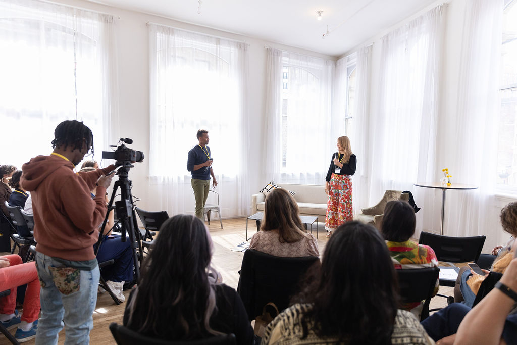 Andre Essue speaking to a seated group in a daylit room while a videographer records the session.